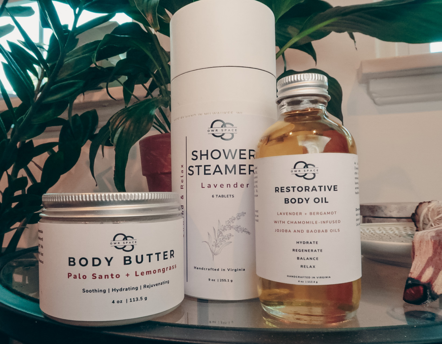 body butter, shower steamer, and body oil aligned together in front of plant during golden hour